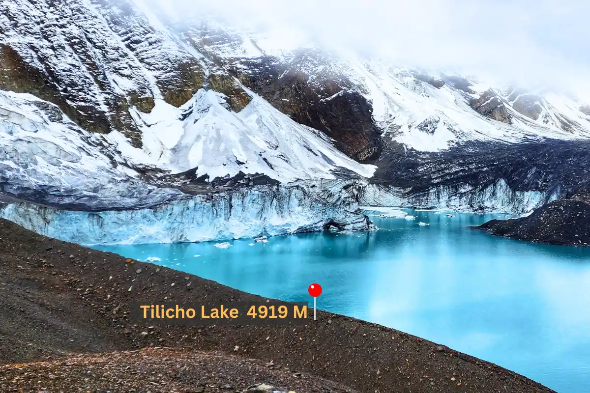 A breathtaking panoramic view of Tilicho Lake, a turquoise glacial lake at 4919 meters, surrounded by snow-capped mountains and dramatic glaciers under a misty sky. A red marker indicates the lake's location.